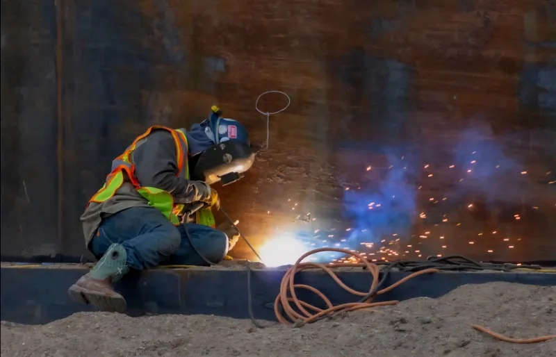 A technician in a hard hat and safety vest welds steel at a mine site. Sparks spray across the ground.